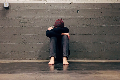 picture of a depressed young man sitting down against a grey brick wall with knees drawn up and his head on his knees